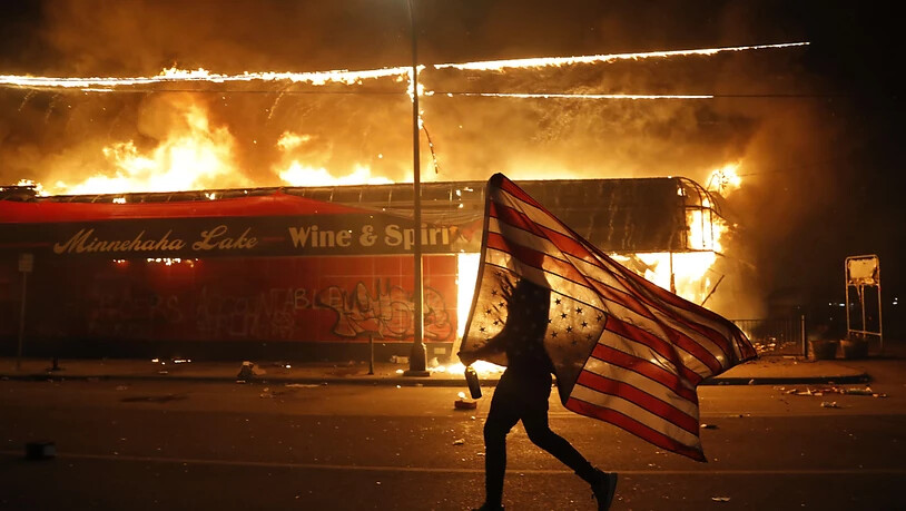 Ein Demonstrant vor einem brennenden Gebäude mit einer eine US-Flagge. Foto: Julio Cortez/AP/dpa