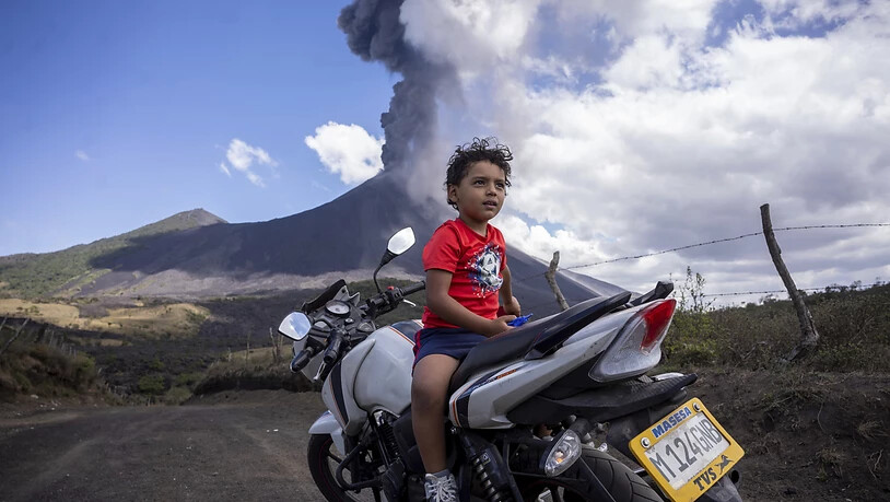 Ein Kind sitzt auf einem Motorrad, während im Hintergrund der Vulkan Pacaya ausbricht. Zum zweiten Mal innerhalb von drei Tagen ist der Vulkan Pacaya in Guatemala ausgebrochen. Foto: Santiago Billy/AP/dpa