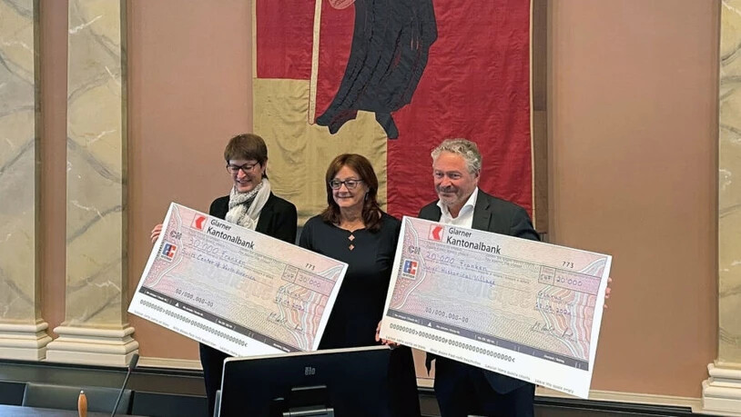 Frau Landamman Marianne Lienhard (links) und Regierungsrat Andrea Bettiga übergeben zwei Checks an Beth Zurbuchen, Präsidentin des Swiss Center of North America.